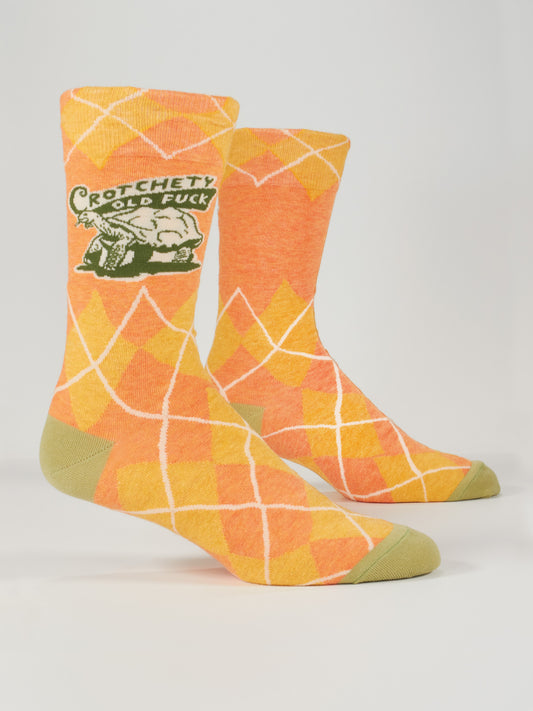 Blue Q’s Crotchety Old Fuck M-Crew Socks feature orange and yellow argyle, green toe and heel accents, plus a cartoon elderly man with “Crotchety Old Fuck” in green. Displayed upright.