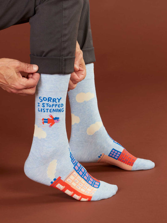 A person wears Blue Q's "Sorry I Stopped Listening" men's socks featuring light blue combed cotton, clouds, buildings, a bird, and the phrase in blue. Fits men's shoe size 7-12. Solid brown background.
