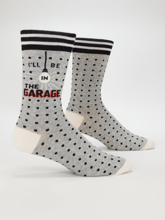 Blue Q’s I'll Be In The Garage M-Crew Socks are gray men’s socks with black polka dots, white toes and heels, black-striped cuffs, a gearshift graphic, and “I’LL BE IN THE GARAGE” text—fun novelty gifts for garage lovers.