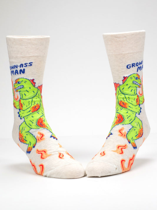 The Blue Q Grown-Ass Man M-Crew Socks are cream-colored novelty socks adorned with a green cartoon monster breathing orange fire, and the phrase “GROWN-ASS MAN” written in blue near the cuff. These funny office socks also feature illustrated orange flames near the toes for an extra-bold statement.