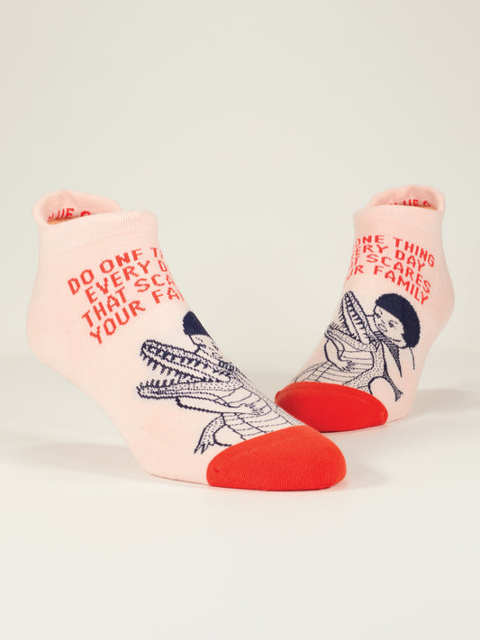 Blue Q's Do One Thing Every Day That Scares Your Family Sneaker Socks are light pink with red toes, feature a person and alligator illustration, and display the bold red text: "Do one thing every day that scares your family.