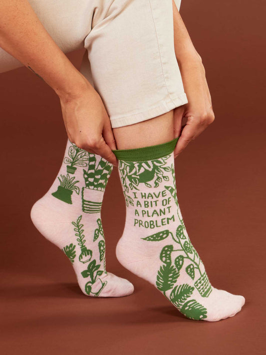 A person in cream pants pulls up Blue Q’s “I Have A Bit Of A Plant Problem” Crew Socks—soft combed cotton socks featuring green potted plants and witty text. Fits women’s shoe size 5-10, shown on a brown background.