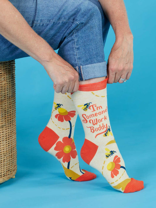 Wearing Blue Q's "I'm Someone's Work Buddy" crew socks, a person in jeans sits on a wicker stool. The colorful, floral cotton socks—size 5-10 for women—feature bees and the phrase, all set against a blue background.