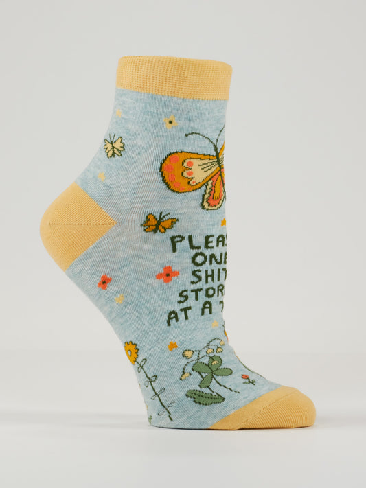 Blue Q’s "Please, One Shitstorm At A Time" W-Ankle Socks are light blue combed cotton with a yellow toe and heel, featuring colorful flowers, a bold orange butterfly, and the cheeky phrase "Please, One Shitstorm At A Time.