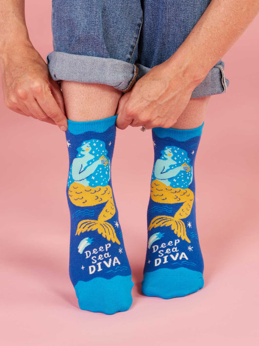 A person adjusts their Blue Q Deep Sea Diva Ankle Socks—blue combed cotton with yellow mermaids and the phrase "Deep Sea Diva"—while standing on a pink surface, jeans rolled up above women's shoes for a fun pop music vibe.