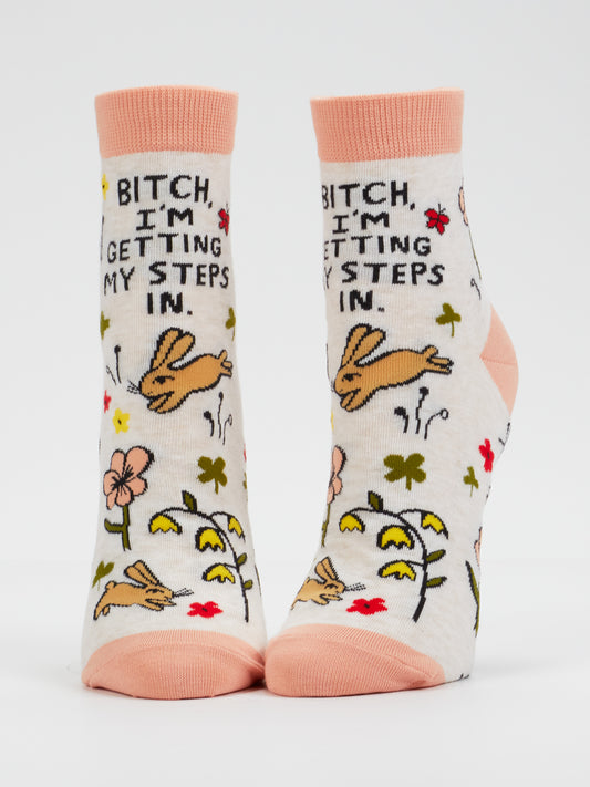 The Blue Q “Bitch, I’m Getting My Steps In” women’s ankle socks feature bunnies, flowers, and the phrase “Bitch, I’m getting my steps in.” in black text on white combed cotton with peach toes and cuffs. Sized to fit women’s shoe sizes 5–10, these socks are shown standing upright against a plain white background.