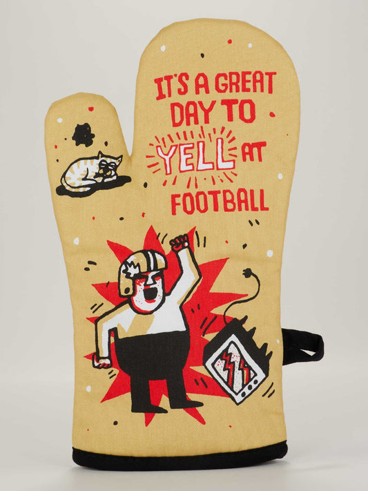 The Blue Q It's A Great Day To Yell At Football Oven Mitt is tan, with a cartoon of someone yelling at a broken TV, a calm cat, and bold red and black text: "IT'S A GREAT DAY TO YELL AT FOOTBALL.
