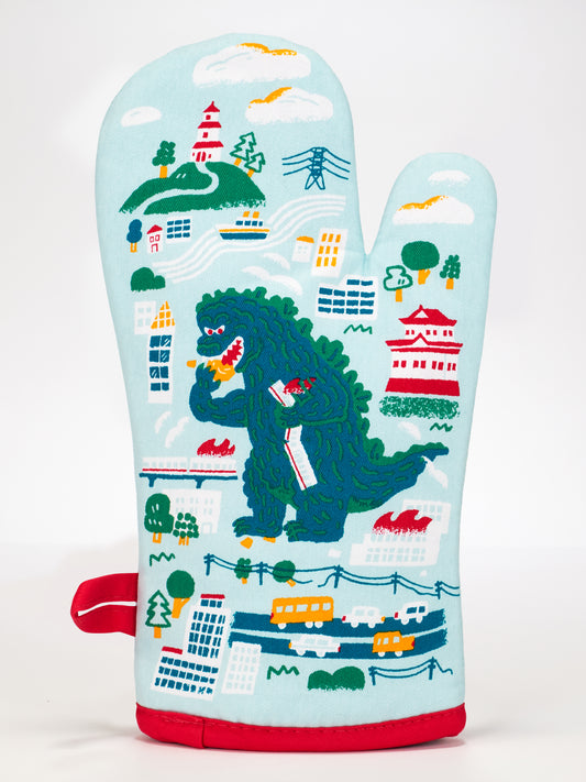 The Blue Q “Fuck Yeah, I Made Cookies.” Oven Mitt is light blue, 100% cotton, features a colorful Godzilla cartoon in a city scene, and has red trim plus a hanging loop. Super-insulated for handling hot pans or cookies safely.