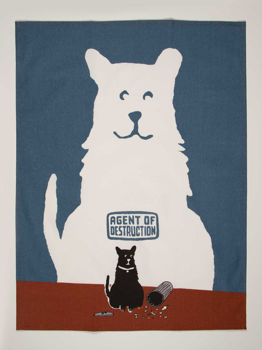 An illustration for Blue Q's "Agent of Destruction Dish Towel" shows a large white dog, tagged as the Agent of Destruction, gently scolding a small black cat next to a spilled trash can and scattered garbage on a table.