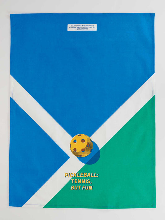The Blue Q "Pickleball: Tennis, But Fun" Dish Towel features a blue, white, and green court with a yellow pickleball and playful text. A small white label accents the top center of this family-friendly towel.