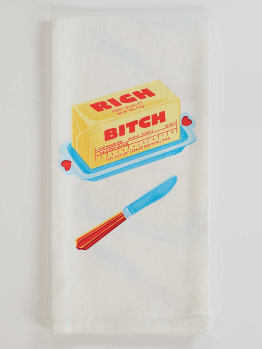 The Blue Q Rich Bitch Dish Towel is a white, unbleached cotton towel featuring a butter stick labeled “Rich Bitch” on a blue dish with red hearts and a red-handled butter knife below.