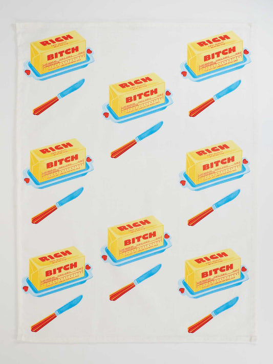 The Rich Bitch Dish Towel by Blue Q is a super-absorbent, unbleached cotton kitchen towel with a diagonal pattern of yellow butter sticks labeled "Rich Bitch Butter," blue dishes, and red-handled butter knives.
