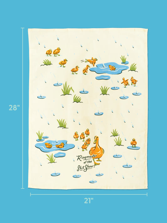 The Blue Q Ringmaster Of The Shitshow Dish Towel is a cream-colored kitchen towel decorated with cartoon ducks, ducklings, lily pads, water drops, and bold “Ringmaster Of The Shitshow” text. Measuring 28 inches by 21 inches, it’s perfect for serving in style and is shown against a blue background.