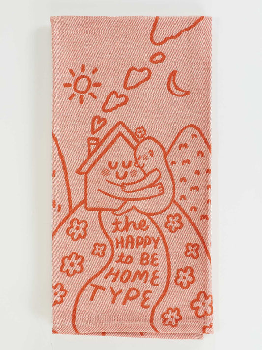 The Happy To Be Home Type Dish Towel by Blue Q features red illustrations of a smiling house, flowers, sun, moon, and hearts on pink cotton, with “The happy to be home type” text. Ideal for those who cherish solitude or calm.