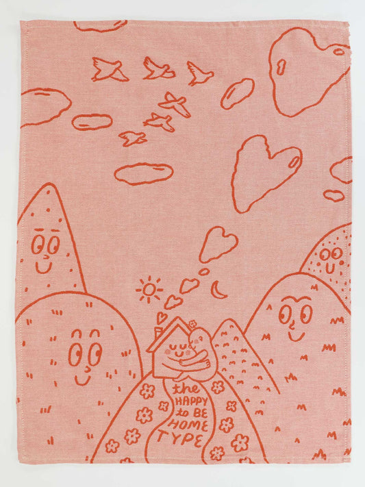 The Blue Q "The Happy To Be Home Type" dish towel is pink with mountains, smiling faces, birds, heart-shaped clouds, and a house with a cat on a hill featuring its namesake phrase.