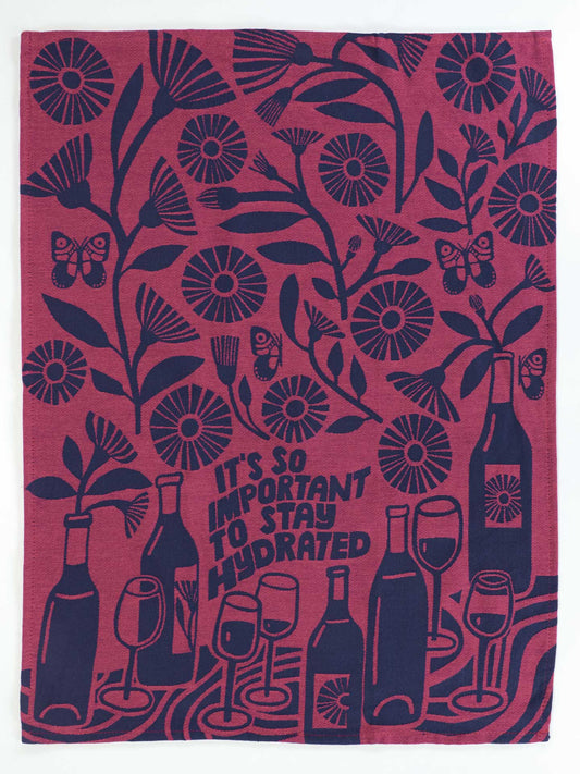 The Blue Q "It's So Important To Stay Hydrated Dish Towel" features dark blue floral designs, butterflies, wine bottles, and glasses on a reddish background with the text: It's so important to stay hydrated for good hydration.