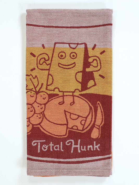 The Blue Q Total Hunk Dish Towel is super-absorbent and shows a cartoon cheese block with arms, flexing on cheese wheels beside grapes. The phrase "Total Hunk" appears at the bottom.