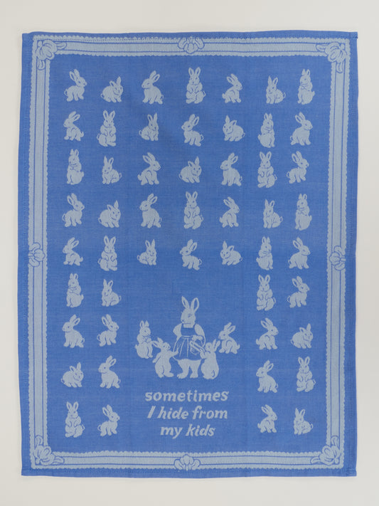 The Blue Q "Sometimes I Hide From My Kids" dish towel is blue, features white illustrations of many small rabbits and a larger rabbit with three smaller bunnies, and is made from unbleached cotton. The text reads: sometimes I hide from my kids.