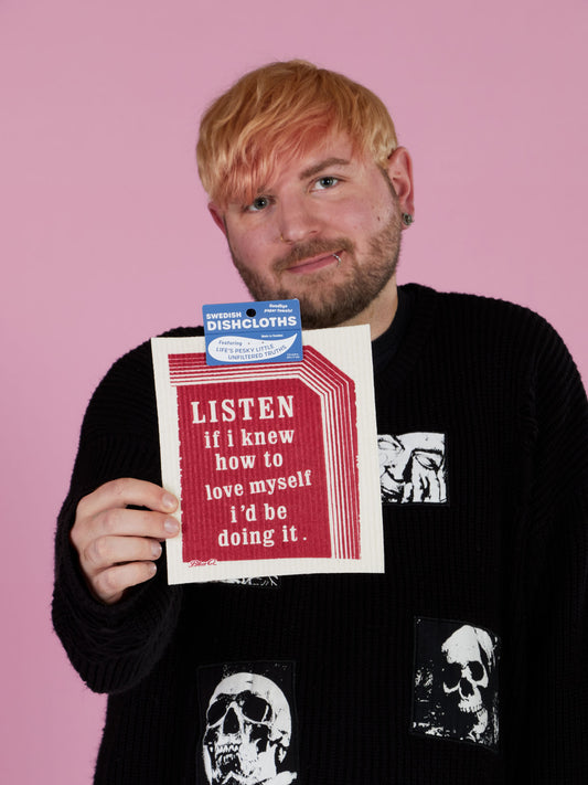 A blond person in a black skull sweater holds up the Blue Q “Listen If I Knew How To Love Myself I'd Be Doing It” Swedish Dishcloth against a pink background.