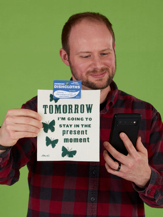 A man in a red plaid shirt smiles while holding a phone and the Blue Q “Tomorrow I’m Going To Stay In The Present Moment” Swedish Dishcloth, against a green background.