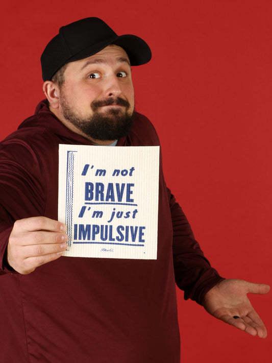 A man in a maroon hoodie and black cap shrugs while holding Blue Q’s "I'm Not Brave I'm Just Impulsive" Swedish Dishcloth against a bold red backdrop—almost as striking as this super-absorbent cloth tackling any mess.