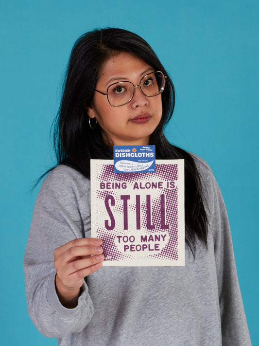 A woman with long dark hair and glasses, in a gray sweatshirt, holds up the Blue Q "Being Alone Is Still Too Many People" Swedish Dishcloth against a plain blue background.