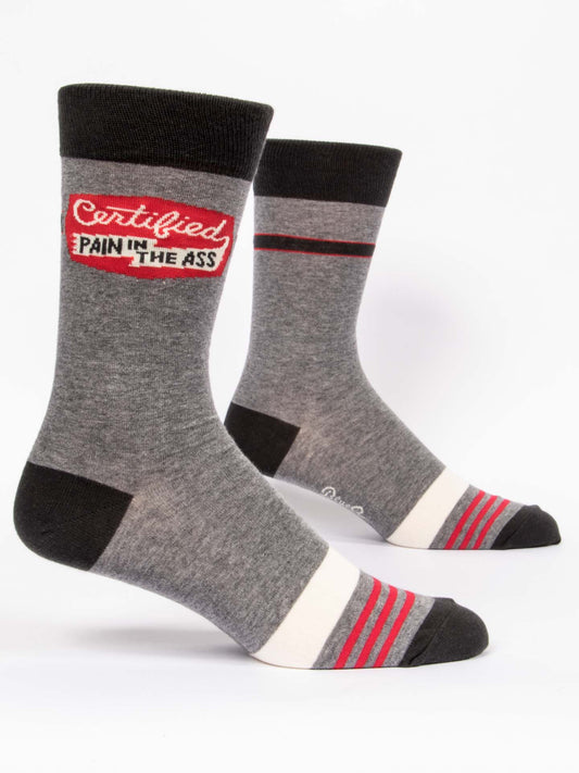 Blue Q’s Certified Pain In The Ass M-Crew Socks are gray combed cotton with black toes, heels, and cuffs, red and white stripes near the foot, and a “Certified Pain in the Ass” patch on each outer side in red, white, and black.