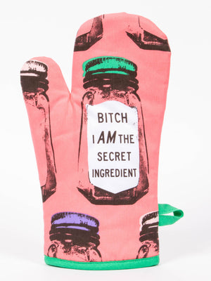 The Blue Q "Bitch I Am The Secret Ingredient" Oven Mitt features pink fabric decorated with spice jars—some labeled paprika, others saffron—and a bold statement on the label. A green fabric loop makes hanging easy.