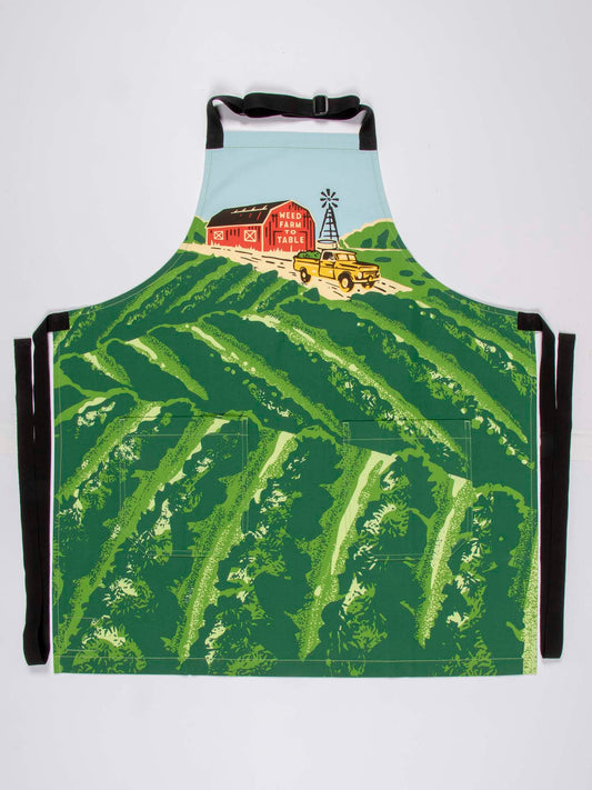The Weed Farm To Table Apron by Blue Q features a vibrant scene of green fields, a red Meal Train barn, a yellow truck, and windmill under blue skies. Adjustable neck strap and black extra-long waist ties provide the perfect fit.