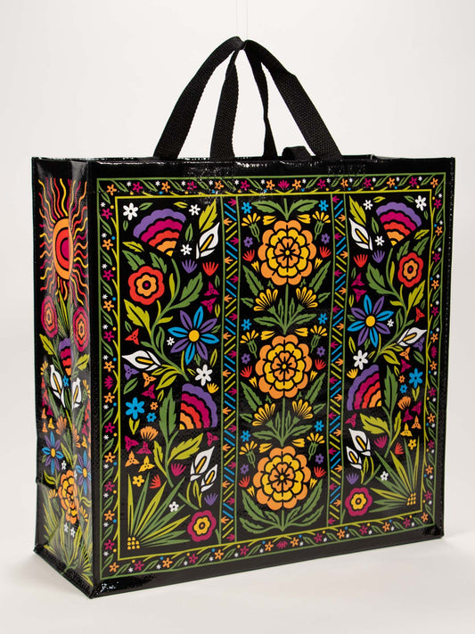 The Blue Q Flower Fest Shopper is a reusable party bag with black handles, featuring vibrant folkloric floral patterns—yellow, orange, red, blue, and white flowers with green leaves on a black background.