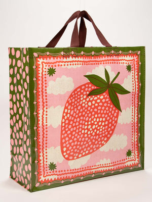 The Blue Q Strawberry Clouds Shopper is a reusable bag with brown handles, green patterned sides, and a whimsical design featuring a large stylized strawberry, white dots, green leaves, and pink background with decorative strawberry clouds.