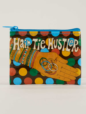 The Blue Q Hair Tie Hustler Coin Purse features a colorful design of a hand holding hair ties, surrounded by purple and yellow polka dots, with "Hair Tie Hustler" in bold, playful lettering.