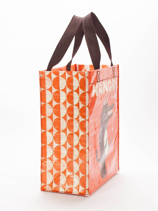 The Blue Q Hangry Handy Tote is an orange reusable bag with brown handles, featuring a geometric white pattern on one side and a “HANGRY” grey creature clutching a veggie burrito on the other.