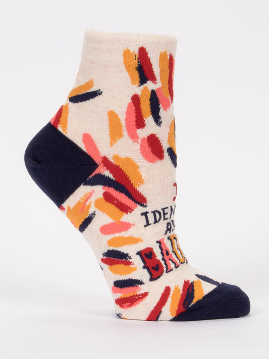 The Blue Q "I Identify As A Badass W-Ankle Socks" are cream-colored with navy toe and heel, abstract red, orange, and yellow brushstrokes, and partial text on the side. Made from soft combed cotton for comfort and style.