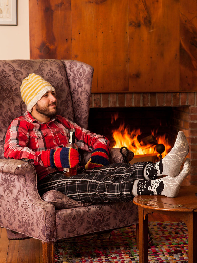 A man in plaid pajamas, colorful mittens, and a striped beanie relaxes by the fireplace, feet up on the table in Blue Q’s Worst Gift Ever M-Crew Socks, holding a drink. The cozy room glows with warmth and comfort.