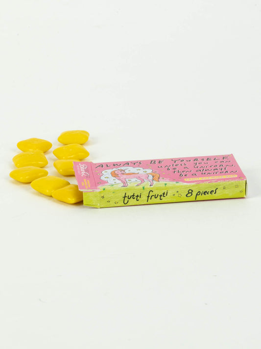 A rectangular Blue Q "Always Be Yourself Unless You Can Be A Unicorn Gum" box with a unicorn illustration is partially open, revealing yellow, heart-shaped candy-coated gum pieces spilling onto a white surface.