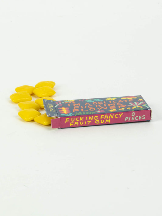 A small purple box labeled "I'm A Delicate Fucking Flower Gum" by Blue Q, 8 pieces, with several yellow fruit-flavored gum pieces scattered beside it on a white background.
