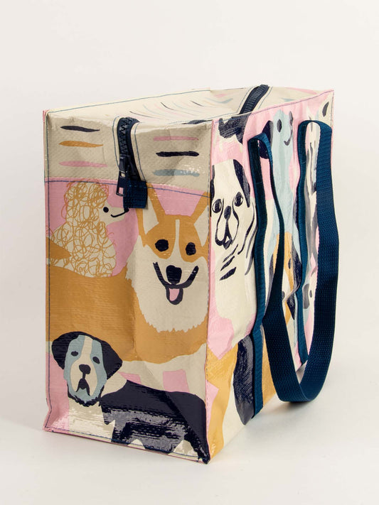 The Blue Q Happy Dogs Shoulder Tote is a sturdy, rectangular reusable bag with blue handles and a zippered top, featuring colorful illustrations of corgis and poodles on a light background.