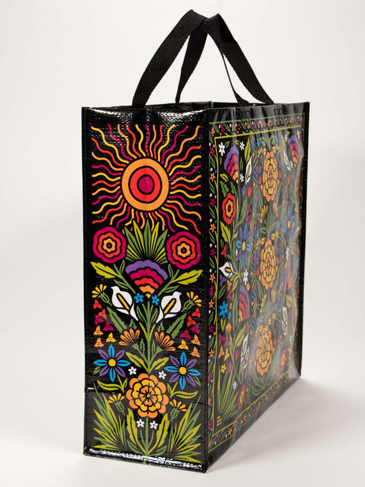 The Blue Q Flower Fest Shopper is a reusable party bag with black handles, displaying vibrant red, orange, yellow, pink, blue, and green floral and sun designs on a glossy black background.