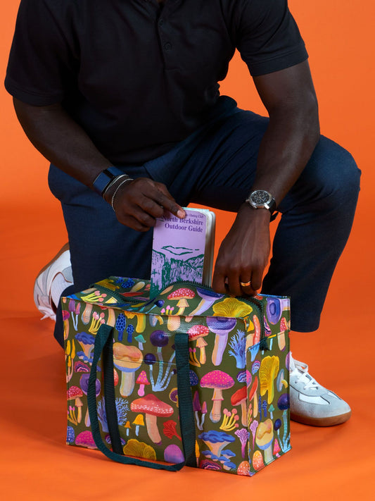 A person kneels on one knee, holding a Wild Berkshire Outdoor Guide book and a Blue Q Mushrooms Shoulder Tote—a reusable bag with a mushroom pattern—against an orange background.