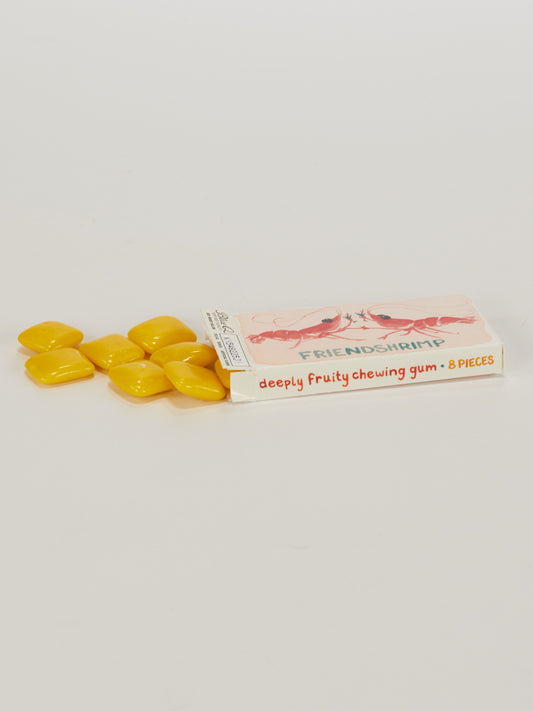 An open pink box labeled Blue Q Friendshrimp Gum – 8 pieces – rests on a white surface, with seven yellow, candy-coated gum pieces spilling out beside it.