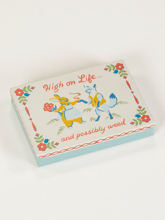 The Blue Q High On Life...And Possibly Weed Pocket Box features cartoon animals—a rabbit and a fox with flowers—and playful red and green floral designs for a cheerful vibe.