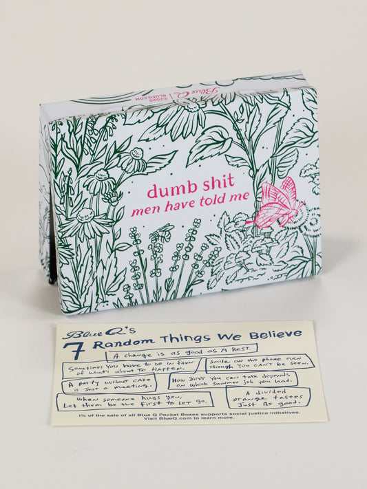 The Blue Q Dumb Shit Men Have Told Me Pocket Box, featuring floral and butterfly art, sits behind a "7 Random Things We Believe" card on a light surface.