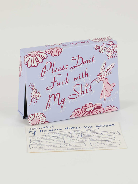 A Blue Q Please Don't Fuck With My Shit Pocket Box, decorated with lavender, pink flowers, and a fairy in cursive font, sits beside a card titled "Random Things We Believe.
