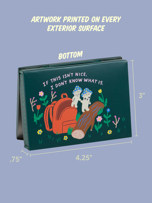 The Souvenirs From My Mushroom Trip Pocket Box by Blue Q is a green rectangular box with artwork of two mushrooms on a log, a red backpack, flowers, pebbles, and "If this isn't nice, I don't know what is" printed above. Box dimensions shown.