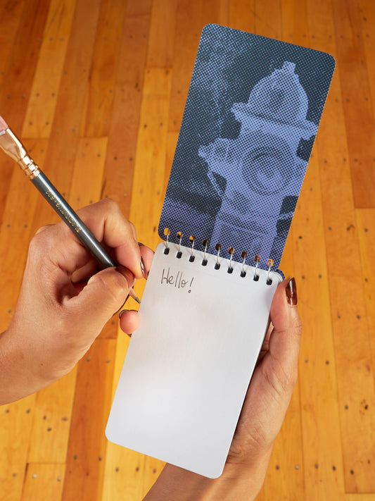Someone writes "Hello!" in pencil on the Blue Q "I Love To Go With The Flow. My Flow, Not Yours" 3x5 spiral notebook, featuring a blue fire hydrant on the cover, with a wooden floor background that complements its design.