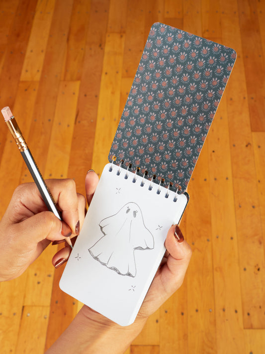 Someone sketches a cartoon ghost in a Blue Q "Ghosts Were People Too" 3x5 Spiral Notebook, its patterned cover visible as it rests above a wooden floor.