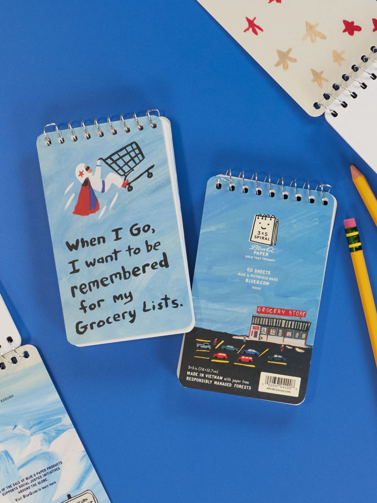 Two Blue Q 3x5 spiral notebooks. One reads "When I Go, I Want To Be Remembered For My Grocery Lists" with a cartoon shopper; the other shows a grocery store illustration on its back cover. Both rest on a blue surface.