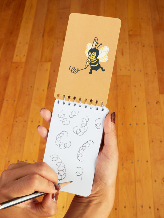On a wooden floor, someone doodles curly loops in the Blue Q Silly Dumb Shit 3x5 Spiral Notebook; above, a flipbook page features a cartoon bee with a pencil drawing silly things.