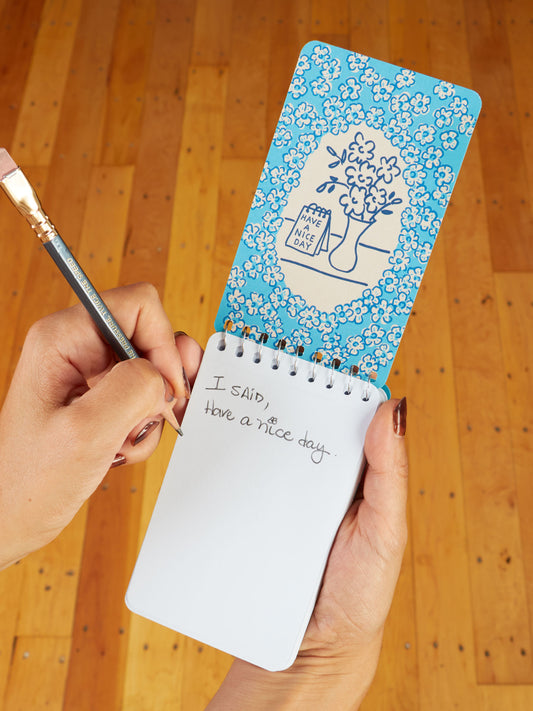 A person writes in a Blue Q Angry Woman Essays 3x5 Spiral Notebook with a pencil. The inside cover has blue florals, a vase, and "have a nice day!".